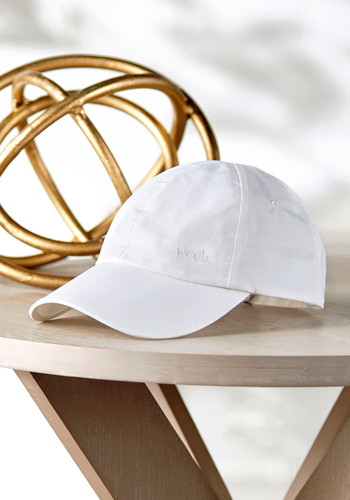 The Woolx Frankie Baseball Hat, featuring "woolx" embroidered on the front, rests on a wooden surface next to a gold sphere. Its white merino wool design and adjustable strap offer functionality, complemented by a softly blurred background for a clean, minimalist look.