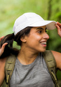 A woman smiles outdoors, wearing the Woolx Frankie Baseball Hat in Polar White, a gray tank top, and a khaki backpack. She holds her ponytail against a lush green backdrop, suggesting she’s hiking or exploring nature.