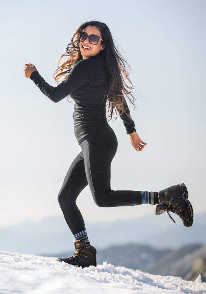 A woman in Woolx Frost Duralite® Leggings—Black, sunglasses, and boots joyfully runs across snowy terrain with mountain views beneath a clear sky.