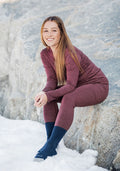 A smiling woman with long brown hair sits on a snowy rock, wearing Woolx Frost Duralite® Leggings in Grape Harvest Melange, a matching long-sleeve top, and dark socks. The rocky backdrop highlights the cold outdoor setting.