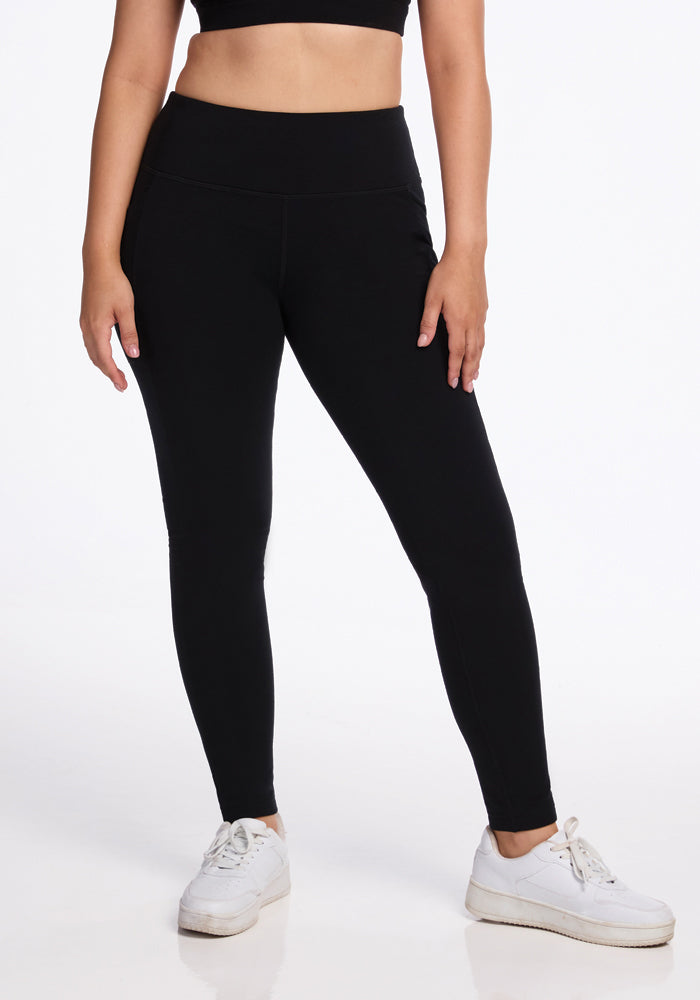 A person stands against a white background, wearing a black crop top, Woolx Frost Duralite® Leggings in black, and white sneakers. The image focuses on their midsection and legs, showcasing a casual and sporty outfit. | Sarah is 5'8", wearing a size S