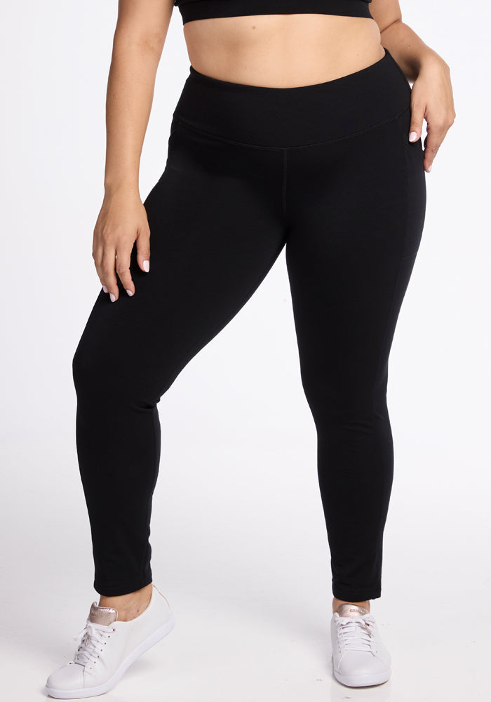 A person with light skin is wearing Woolx's Frost Duralite® Leggings in black paired with a black crop top. They are also wearing white sneakers against a plain white backdrop. The individual stands with one arm hanging loose while the other rests on their hip. | Juliana is 5'10", wearing a size XL