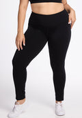 A person with light skin is wearing Woolx's Frost Duralite® Leggings in black paired with a black crop top. They are also wearing white sneakers against a plain white backdrop. The individual stands with one arm hanging loose while the other rests on their hip. | Juliana is 5'10", wearing a size XL