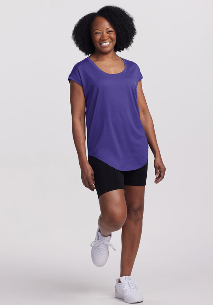 Woman in Woolx Gabi Tunic Top - Berry Indigo and black shorts, wearing white sneakers, stands on one leg.