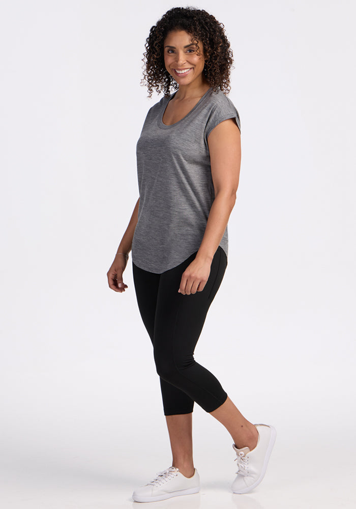 A woman with curly hair smiles while standing sideways, wearing the Woolx Gabi Tunic Top in Graphite Heather, black cropped leggings, and white sneakers against a plain white background.