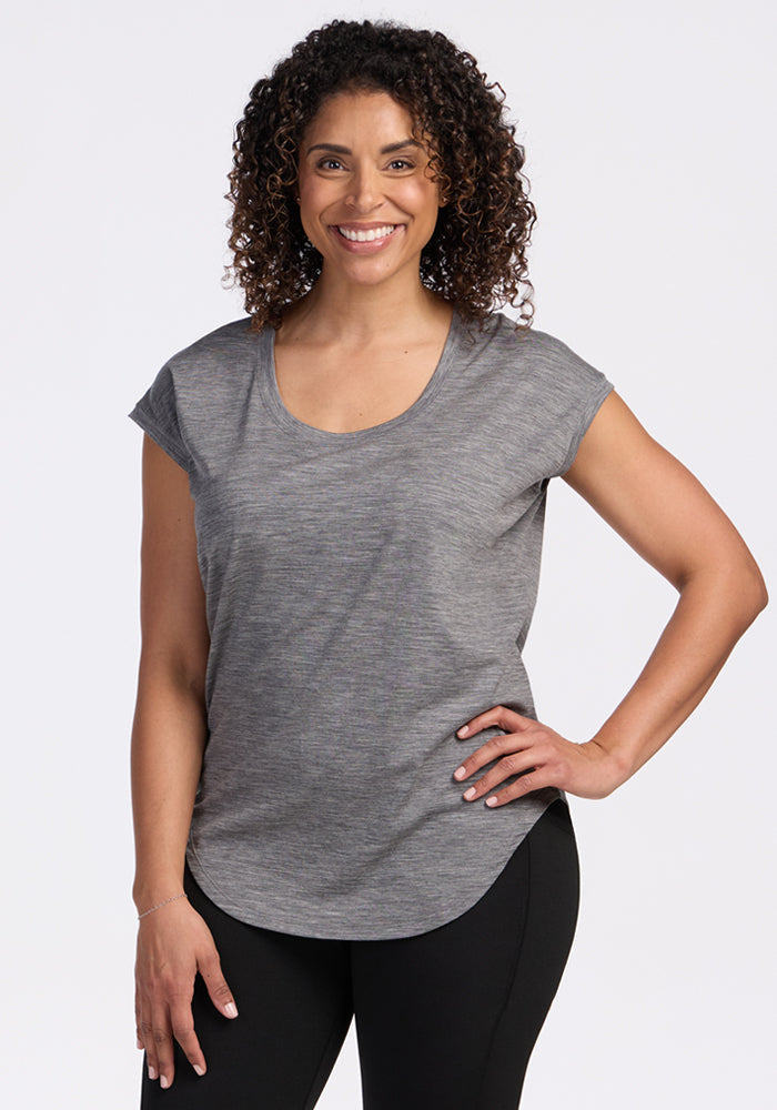 A woman with curly hair smiles at the camera, wearing the Woolx Gabi Tunic Top in Graphite Heather and black pants. She stands with one hand on her hip against a plain, light background. | Kisha is 5’8”, wearing a size M