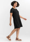 A person with curly hair smiles while moving sideways, wearing a short-sleeved black Georgie Dress by Woolx and brown platform sandals. The white background highlights their confident and relaxed demeanor. | Tori is 5'7", wearing a size S