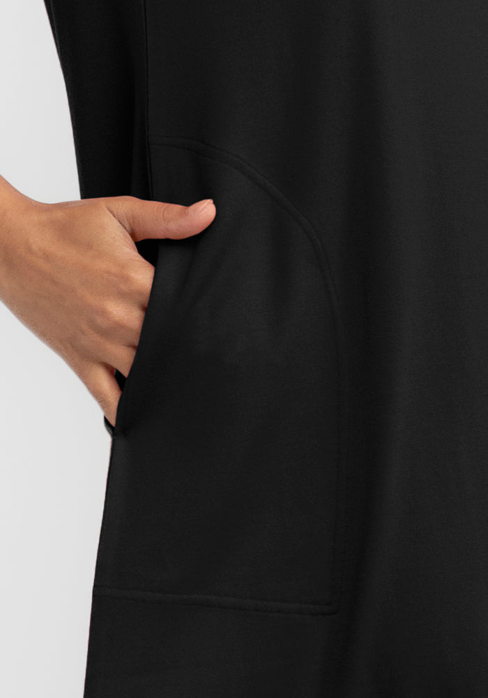 A close-up shows a person wearing the Georgie Dress - Black by Woolx, with their hand in a side pocket. The plain background highlights the simplicity and elegant A-line design of this merino wool garment.