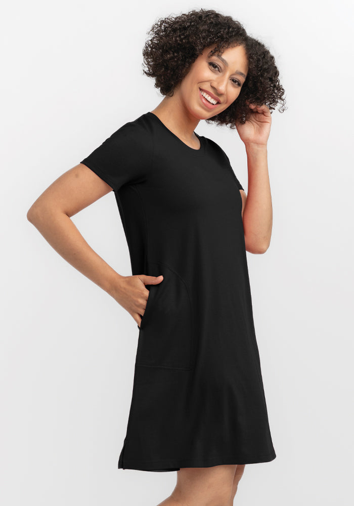 A person with curly hair smiles in a black Woolx Georgie Dress, made of merino wool. They pose with one hand in their hair and the other in a pocket against a plain white background.