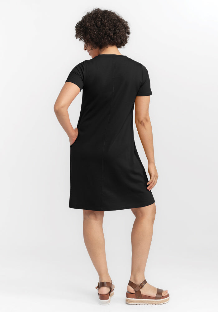 A curly-haired person faces away from the camera, wearing a short-sleeved Woolx Georgie Dress in black with brown platform sandals. The plain white background accentuates the ensemble's sophisticated simplicity.