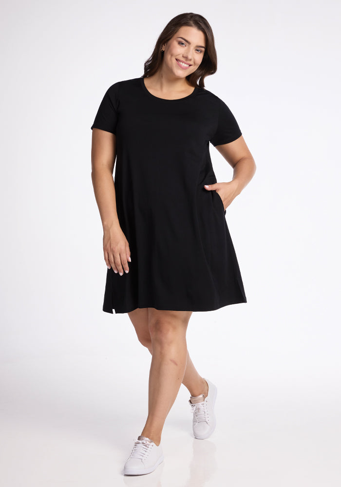 A person smiles, standing against a plain white background, wearing the Georgie Dress - Black by Woolx. This A-line silhouette dress is crafted from merino wool and features short sleeves and pockets. Paired with white sneakers, they have one hand in their dress pocket. | Juliana is 5'10", wearing a size XL