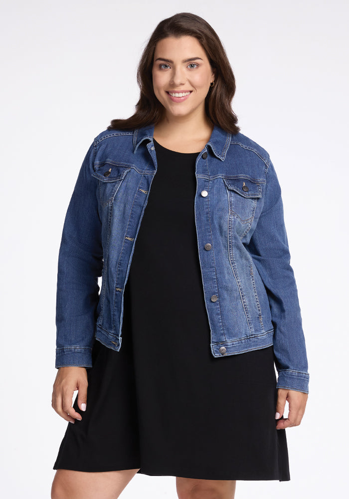 A smiling woman with long brown hair wears a blue denim jacket over a chic Georgie Dress - Black by Woolx, standing against a plain light background.