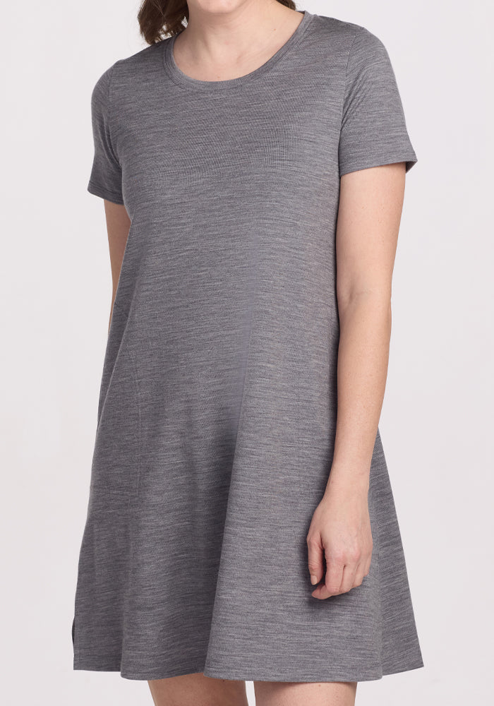 A person wears the Woolx Georgie Dress in Graphite Heather, crafted from cooling Merino wool. Posed against a white background, the short-sleeve dress fans out gracefully at the hem for comfort and elegance while they stand with their gaze averted from the camera.