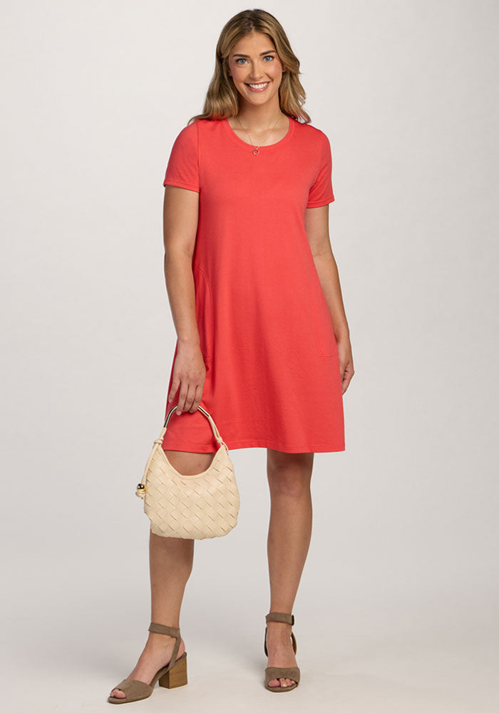 Woman in the Woolx Georgie Dress - Hibiscus holds a cream woven handbag and wears tan block-heel sandals, smiling. | Ariana is 5'11", wearing a size S