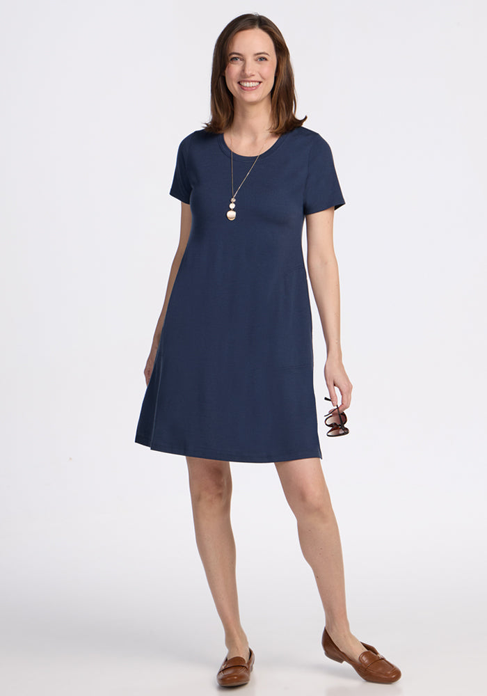 Woman wearing the Woolx Georgie Dress in Deep Navy, brown flats, a necklace, holding eyeglasses, and smiling.