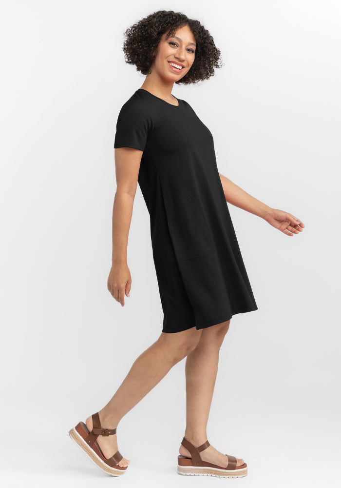 A person with curly hair smiles while moving sideways, wearing a short-sleeved black Georgie Dress by Woolx and brown platform sandals. The white background highlights their confident and relaxed demeanor. | Tori is 5'7", wearing a size S