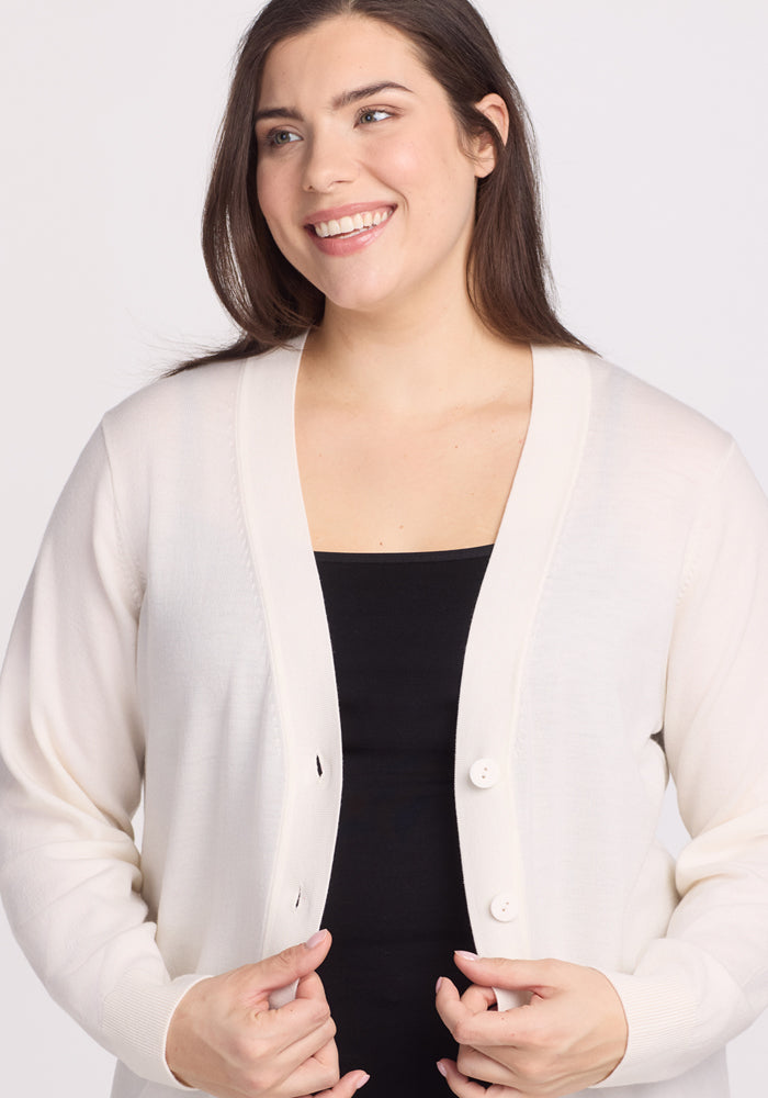 A person with long dark hair smiles, wearing the Woolx Gia Sweater - Dewdrop Pearl, made from moisture-wicking Merino wool, over a black top. The background is plain and light-colored.