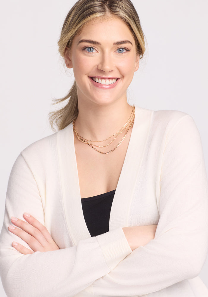 A light-haired, fair-skinned woman crosses her arms and smiles in a serene setting against a plain white background. She wears a Woolx Gia Sweater in Dewdrop Pearl over a black top and layered necklaces, offering lightweight comfort.