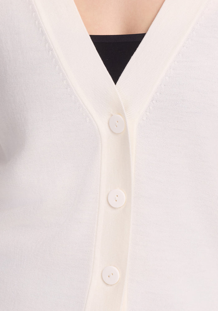 Close-up of the Gia Sweater - Dewdrop Pearl by Woolx, a white cardigan made from odor-resistant Merino wool with a V-neck and four round white buttons. A black top underneath highlights its lightweight comfort and slightly textured, soft fabric.