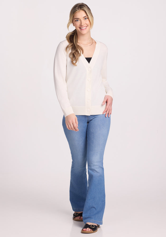 A person with long hair smiles while wearing the Woolx Gia Sweater in Dewdrop Pearl over a black top and light blue jeans. They stand with one hand in their pocket against a plain white background, showcasing effortless style and comfort.