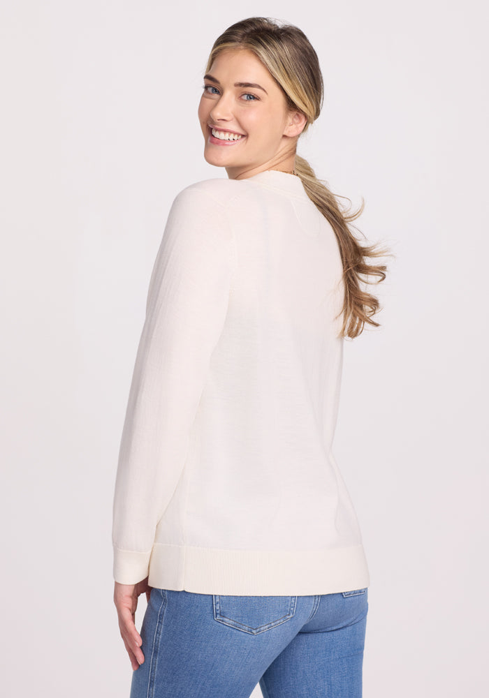 A smiling woman with long hair wears the Gia Sweater - Dewdrop Pearl by Woolx and blue jeans, looking over her shoulder against a plain background.