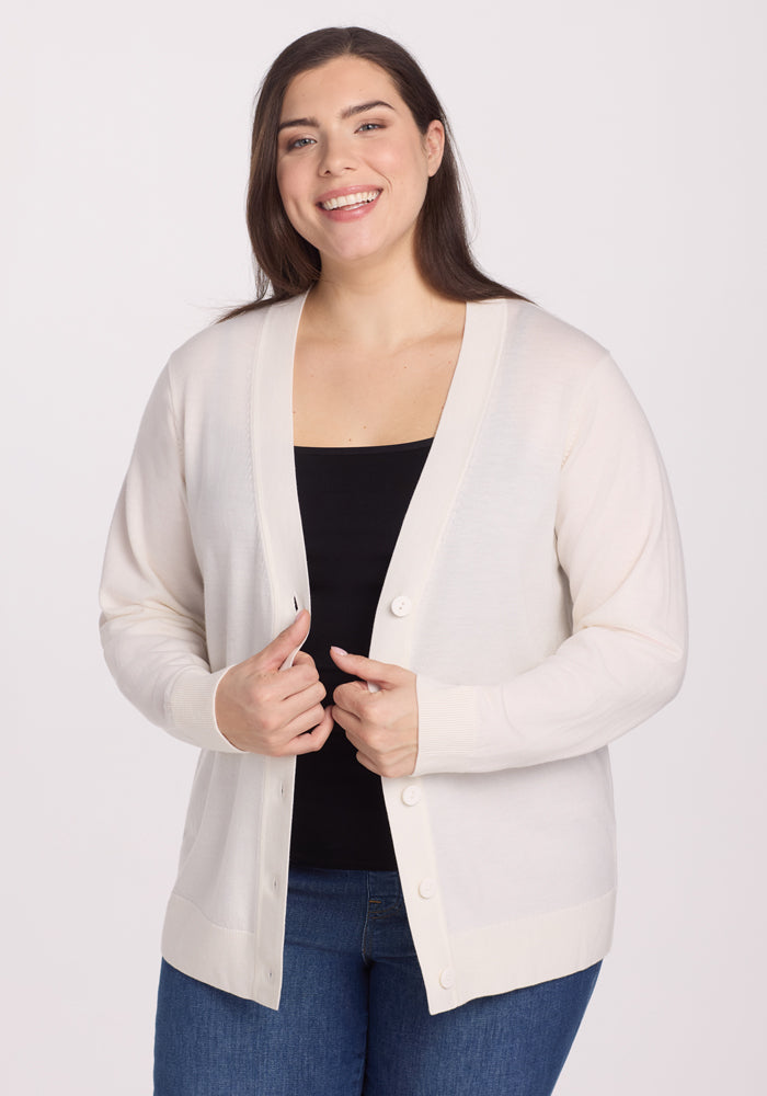A person smiles while wearing the Woolx Gia Sweater in Dewdrop Pearl, made of odor-resistant Merino wool, over a black top and blue jeans. They stand against a plain white background, holding the lightweight cardigan's edges together. | Juliana is 5'10", wearing a size XL