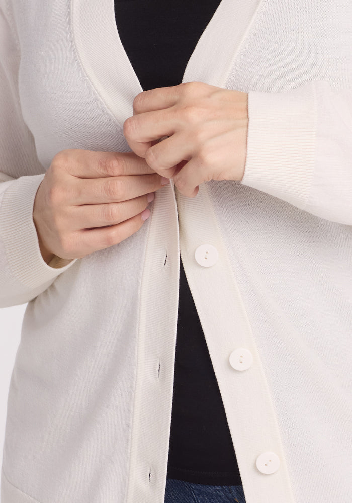 A person is buttoning the Gia Sweater - Dewdrop Pearl, a lightweight, Merino wool cardigan by Woolx with white buttons over a black top, shown from neck to waist on a plain, light background.
