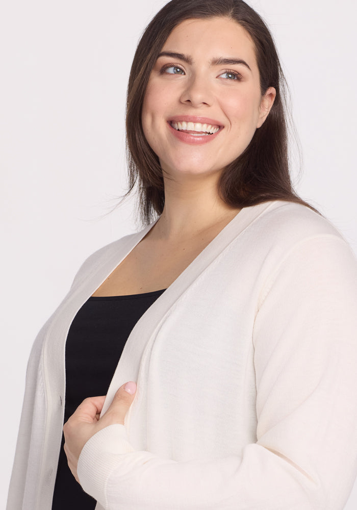 A person with long brown hair smiles warmly while wrapped in the cozy comfort of the Gia Sweater - Dewdrop Pearl by Woolx. They wear it over a black top, glancing to the side against a plain white background, effortlessly blending warmth and style.