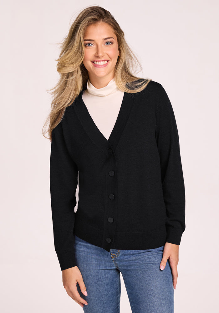 A woman with long blonde hair smiles against a light background, wearing a Woolx Gia Sweater in black over a white turtleneck and blue jeans, enjoying the cozy, odor-resistant comfort of this classic Merino wool style. | Ariana is 5'11", wearing a size S