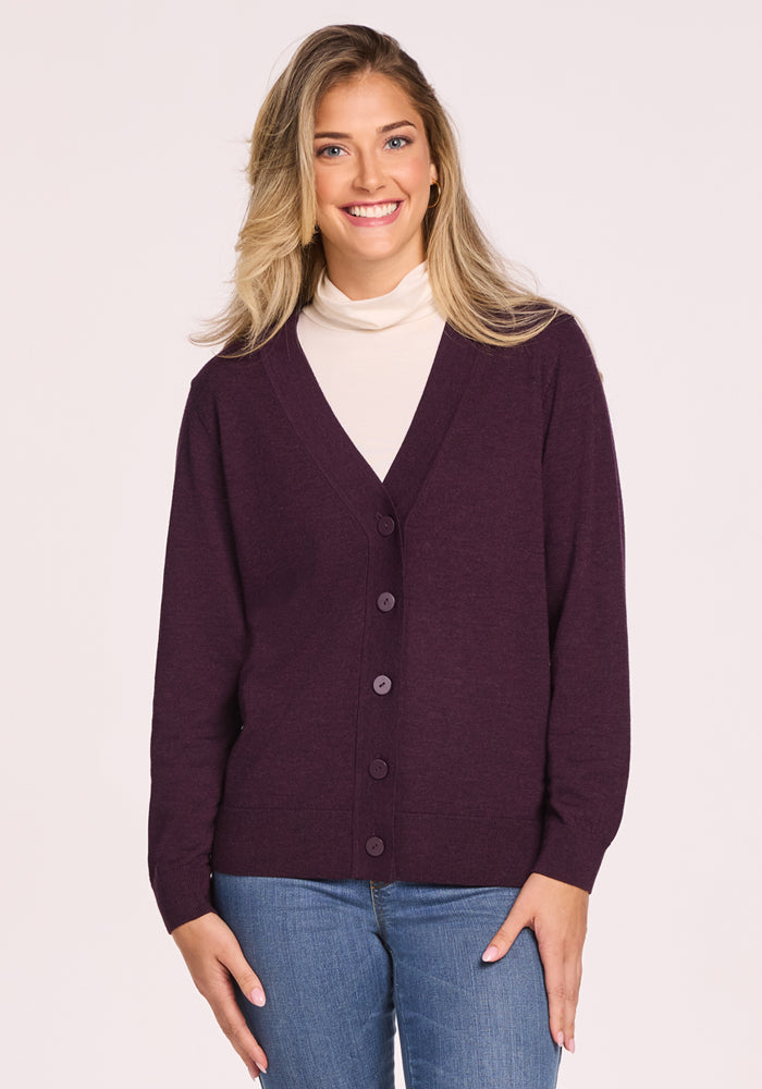 A woman with long blonde hair smiles at the camera, wearing the Woolx Gia Sweater in Deep Plum layered over a cream turtleneck and paired with blue jeans, standing against a light background.