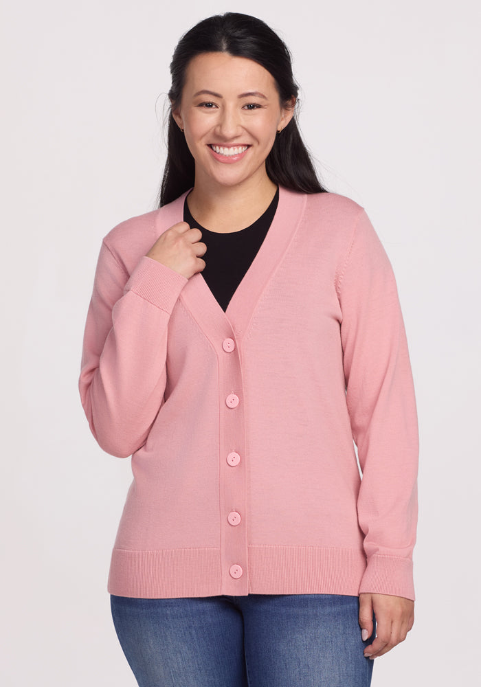 A woman with long dark hair smiles, wearing the Woolx Gia Sweater in Blush over a black top and blue jeans against a light background. This lightweight Merino wool, odor-resistant sweater adds comfort and style to her look.