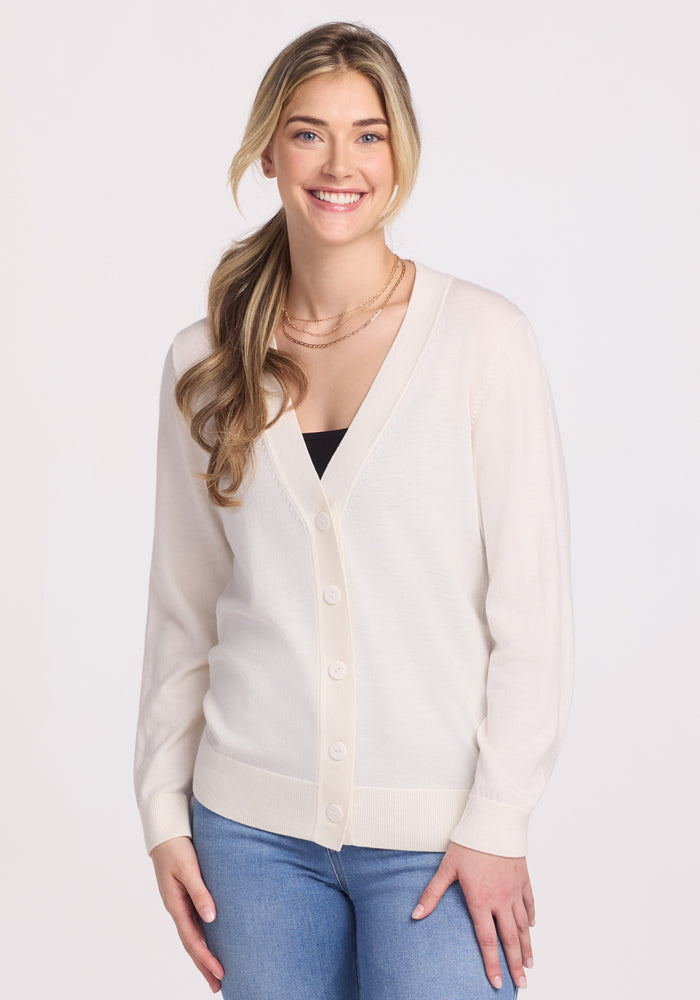 A woman with long blonde hair in a ponytail is wearing the Gia Sweater - Dewdrop Pearl by Woolx over a black top and blue jeans against a plain white background. | Ariana is 5’11”, wearing a size S