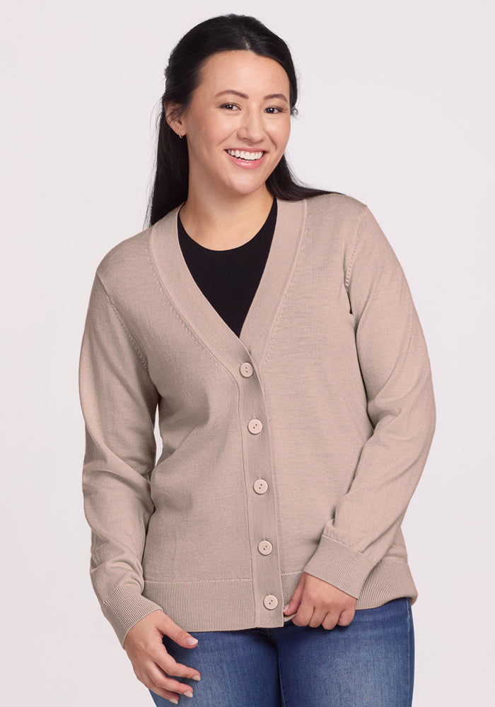 A woman with long dark hair wears the Woolx Gia Sweater in Sandstone Haze over a black top and blue jeans, smiling against a plain light background. Final Sale.