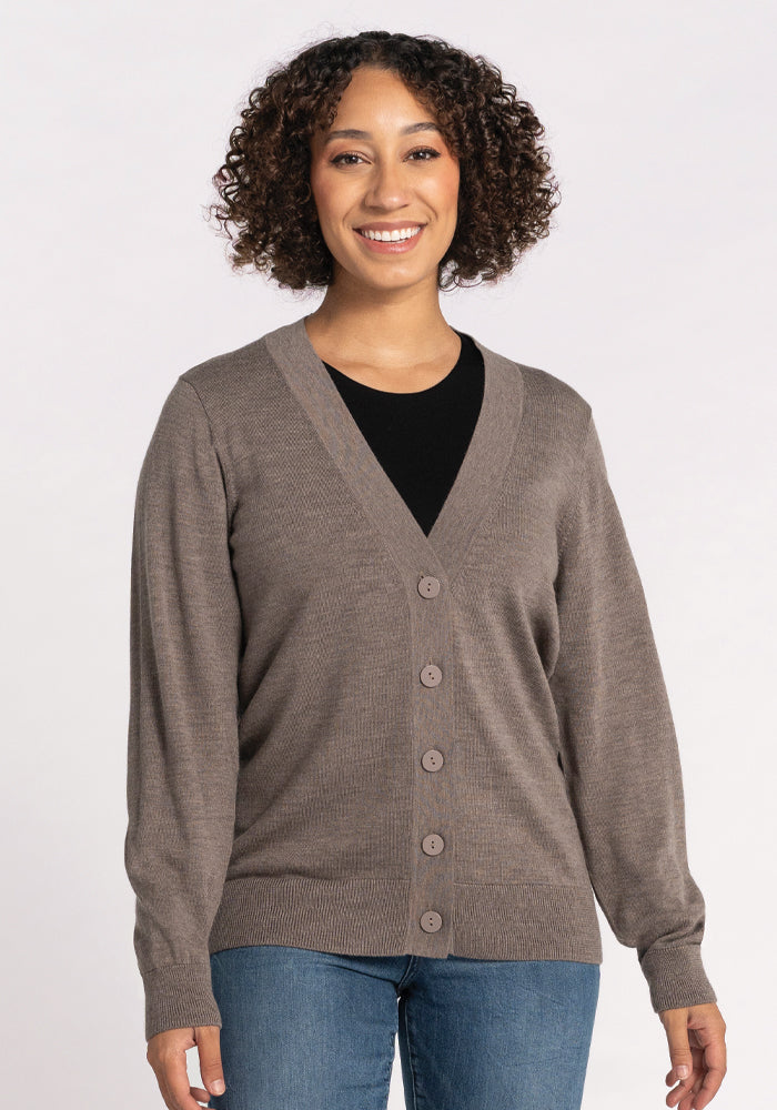A woman with curly hair smiles, wearing the Woolx Gia Sweater in Sable over a black top and blue jeans, standing against a plain light background.