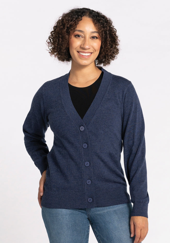 A smiling woman with curly hair wears the Woolx Gia Sweater in Twilight Storm over a black top and blue jeans, standing against a plain light background.