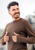 A man with short dark hair and a beard smiles and gives two thumbs up outdoors. He is wearing the Woolx Glacier Long Sleeve Crew • Final Sale - Bourbon Oak.