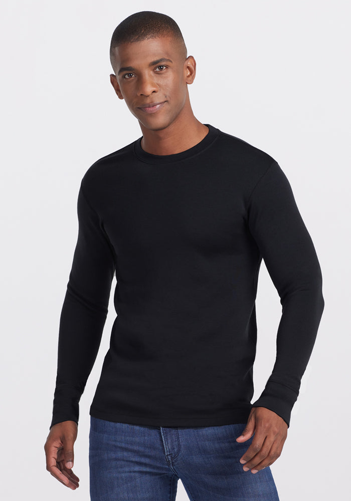 A man wearing the Woolx Glacier Long Sleeve Crew in black and blue jeans stands against a white background, looking at the camera with a slight smile.