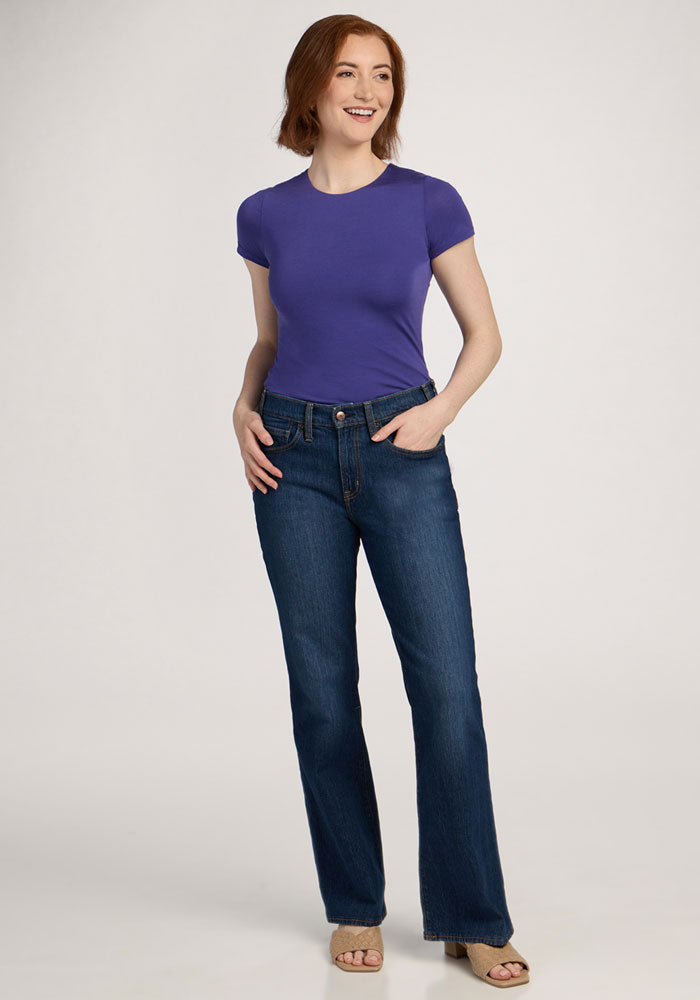 Woman standing and smiling in a Woolx Gracie Bodysuit - Berry Indigo with blue jeans and beige sandals.