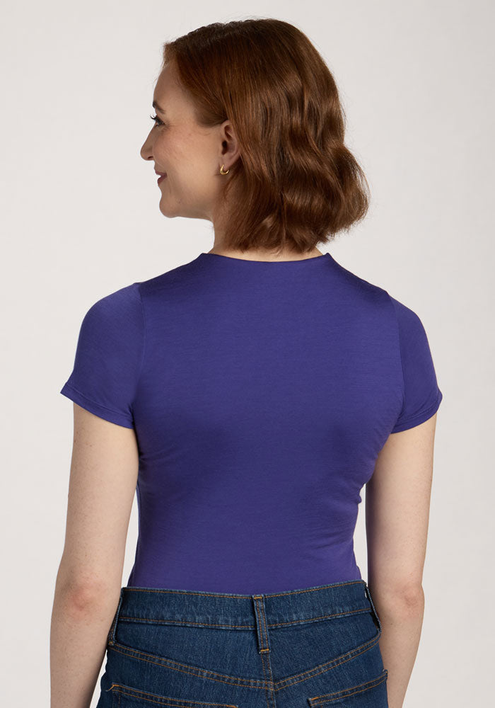 A woman in the Woolx Gracie Bodysuit - Berry Indigo and jeans stands with her back to the camera, looking left.
