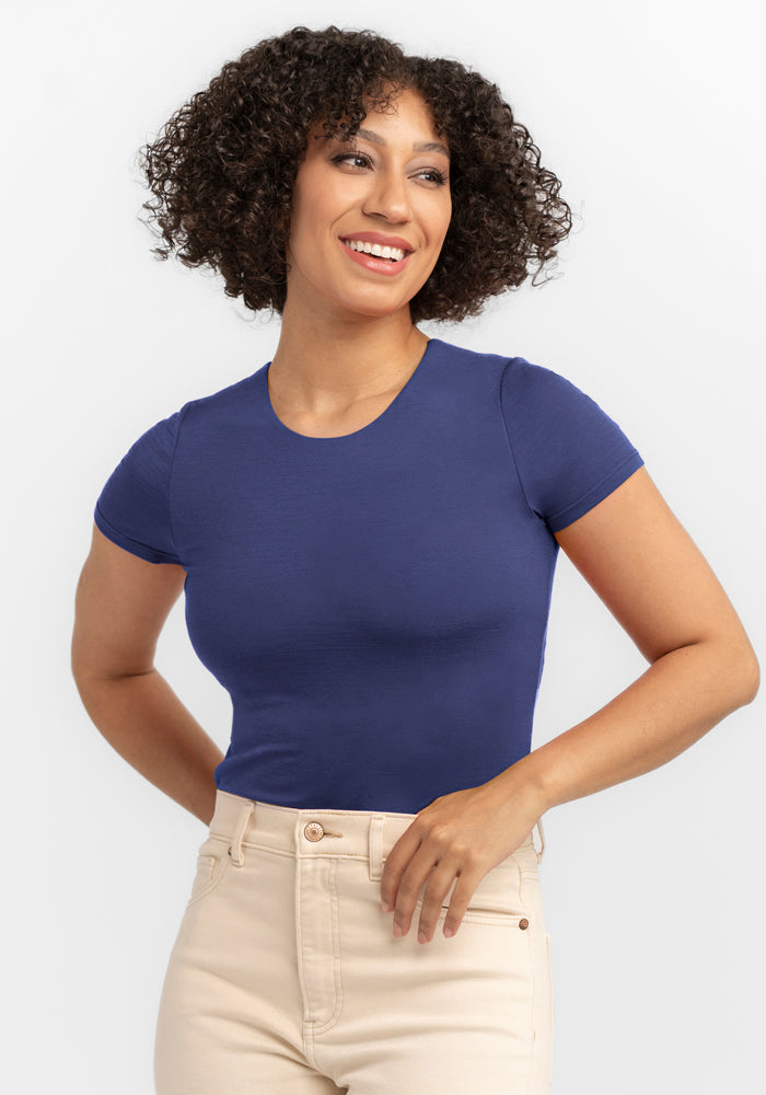 A person with curly hair smiles warmly, wearing the Woolx Gracie Bodysuit in Blue Horizon and beige pants, standing against a plain background. | Tori is 5'7", wearing a size S