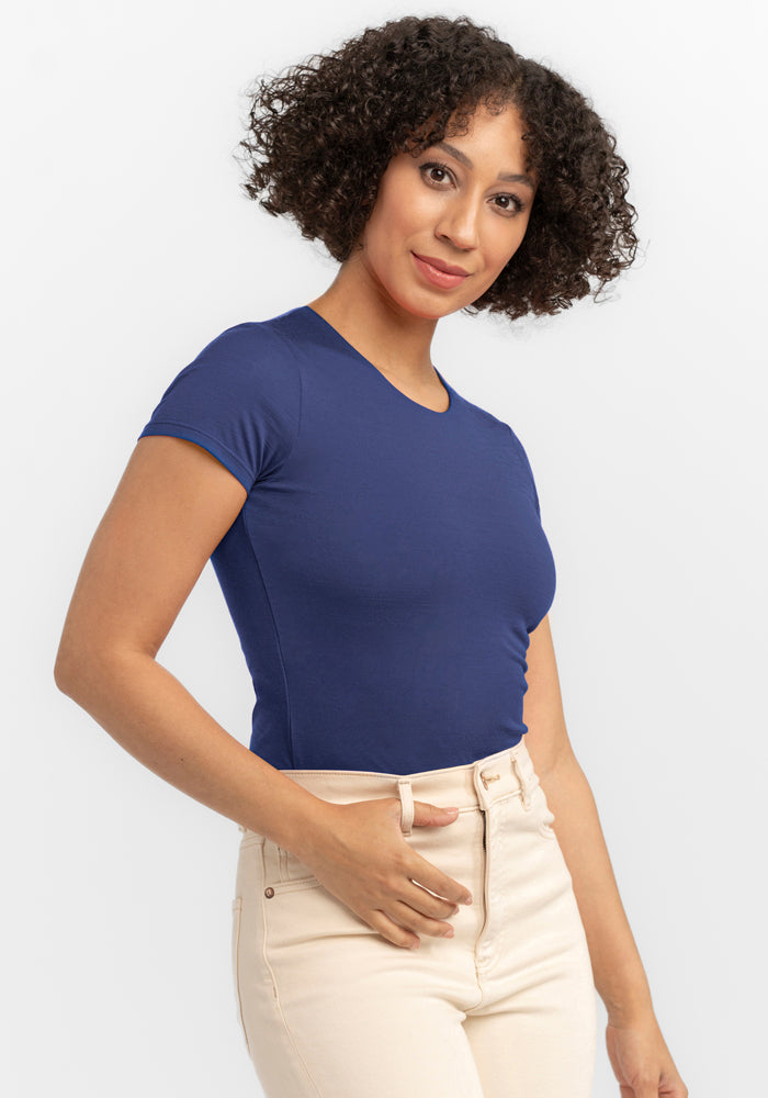 A woman with curly hair models the Woolx Gracie Bodysuit in Blue Horizon, paired with light pants, against a plain white background.