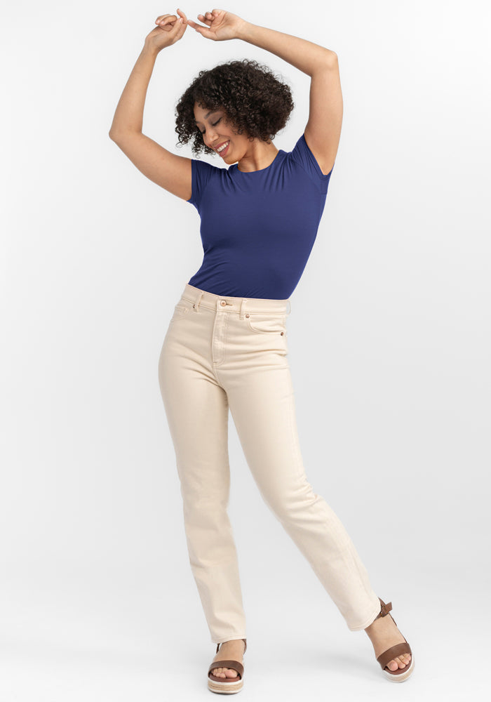 A curly-haired person smiles, posing with raised arms in a temperature-regulating Gracie Bodysuit - Blue Horizon by Woolx. They pair it with beige pants and brown sandals against a plain white background.
