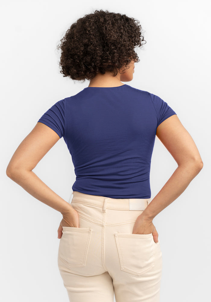 A person with short curly hair faces away, wearing the Woolx Gracie Bodysuit - Blue Horizon and light beige pants. Their hands rest in the back pockets against a plain white backdrop.