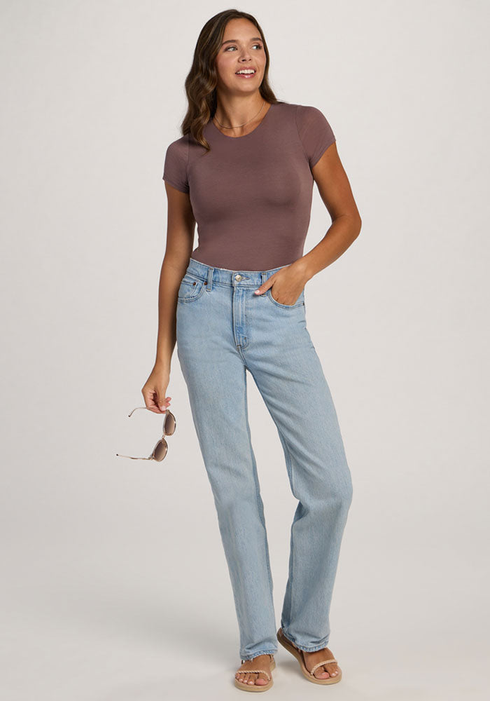 Woman in Woolx Gracie Bodysuit - Peppercorn and light blue jeans smiles, holding sunglasses with one hand in her pocket.