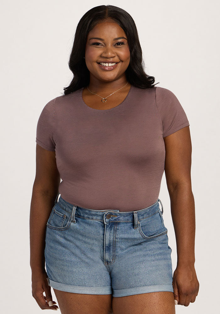 A smiling woman in the Woolx Gracie Bodysuit - Peppercorn stands against a plain, light background. | Sapphire is 5’8”, wearing a size XL