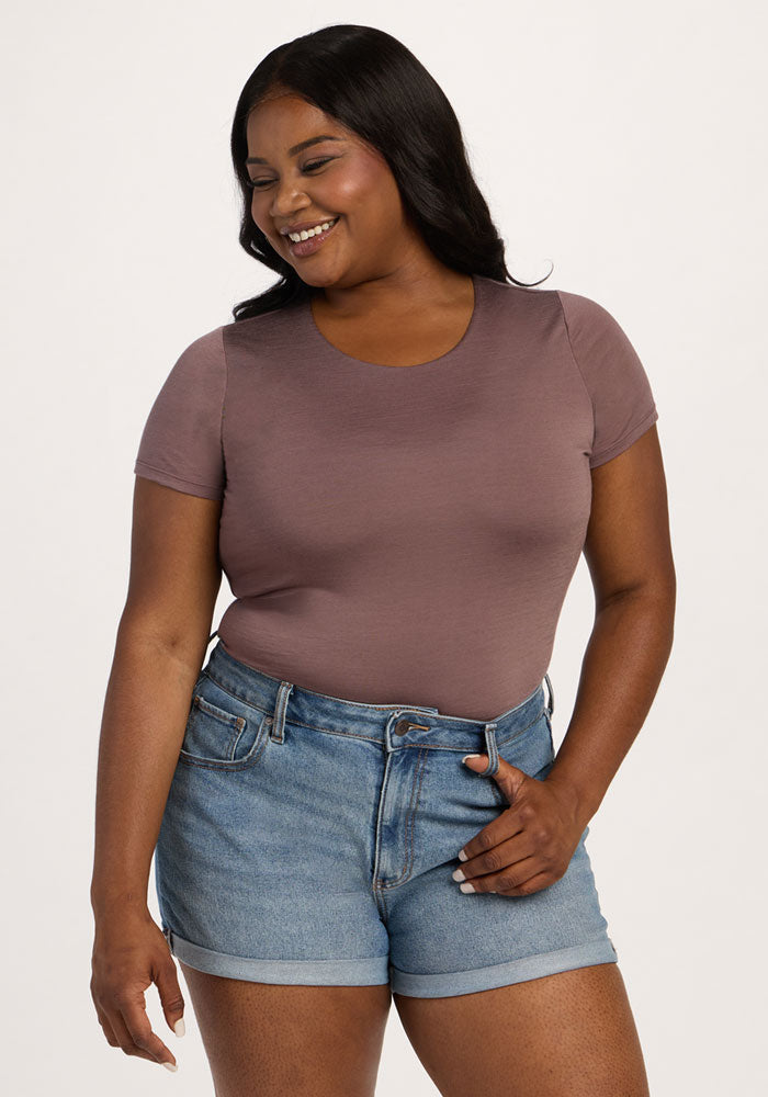 Smiling woman wears the Woolx Gracie Bodysuit in Peppercorn and denim shorts, standing with a hand in her pocket.