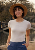A woman wears the Woolx Gracie Bodysuit in Vanilla, paired with blue pants and a tan hat, as she stands outdoors smiling gently in front of a bridge over water, with trees in the background and her hands in her pockets.