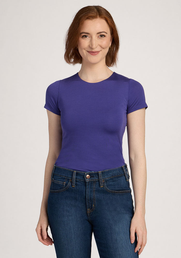 A woman smiles in a Woolx Gracie Bodysuit - Berry Indigo with blue jeans, short auburn hair, and a plain background. | Shea is 5’9”, wearing a size S