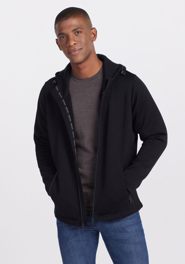 A person stands against a plain background, dressed in a black Woolx Grizzly Sweatshirt over a gray t-shirt and blue jeans. They have short hair and their hands are casually placed in the pockets of the sweatshirt, smiling slightly at the camera. | Trell is 6’2”, wearing a size M