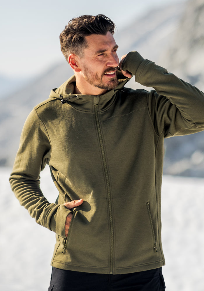 A man with short brown hair and a beard stands outdoors in snowy mountains, wearing the Woolx Grizzly Sweatshirt in black, with his left hand near his mouth and his right hand in his pocket.
